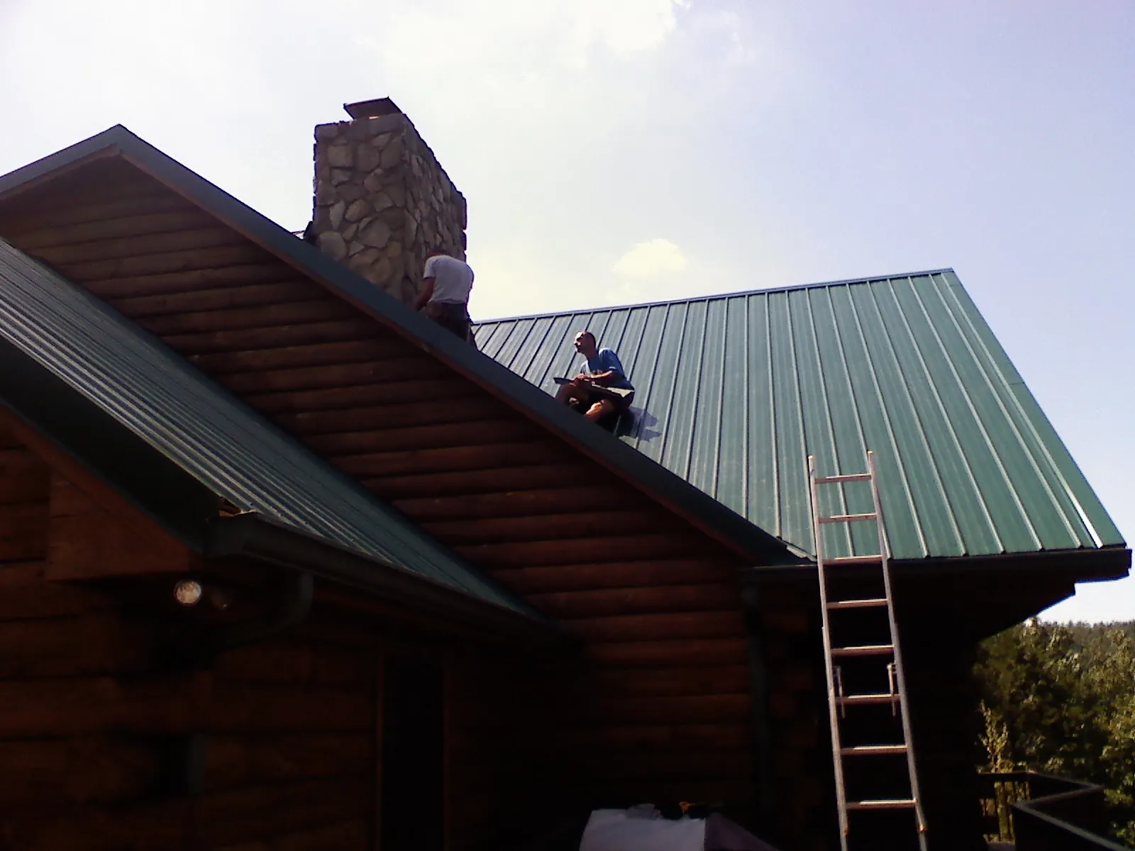 Roofers installing green metal panels beside a stone chimney on a log home in Avoca